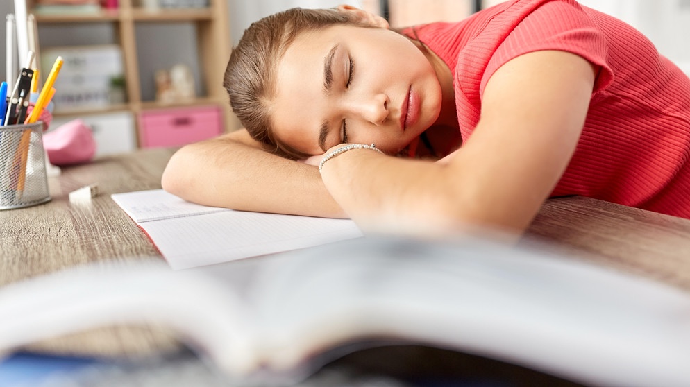Junges Mädchen liegt mit dem Kopf müde auf dem Schreibtisch. Hat die Zeitumstellung gesundheitliche Folgen? Und warum wird überhaupt an der Uhr gedreht? | Bild: picture alliance / Zoonar | lev dolgachov