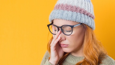 Eine müde wirkende junge Frau mit Brille und Mütze hält sich mit einer Hand die Augen zu. Krisen aushalten und überwinden - und dabei leistungsfähig bleiben. Unzählige Ratgeber behaupten, das ließe sich trainieren. Wie sinnvoll ist das? Setzen wir uns damit nicht erst recht unter Druck? | Bild: colourbox.com